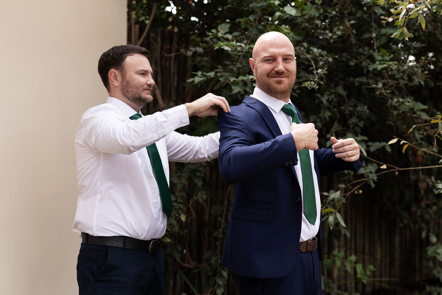 Two men in formal wear helping each other adjust their matching green ties.