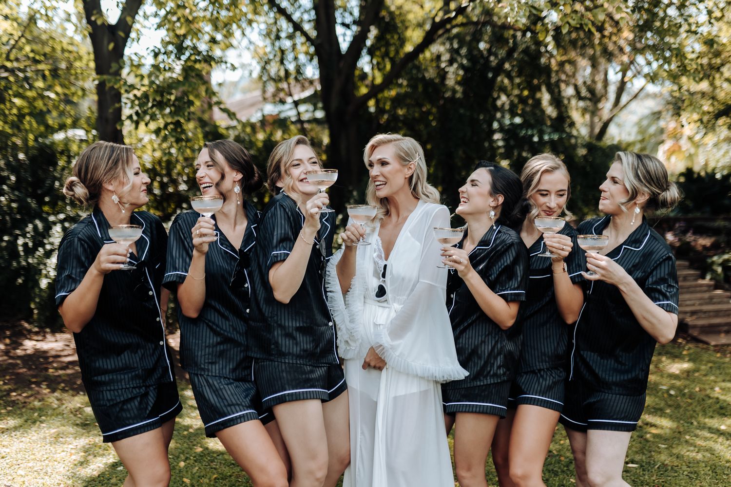 Bride in white robe laughing with bridesmaids in black robes during outdoor celebration.