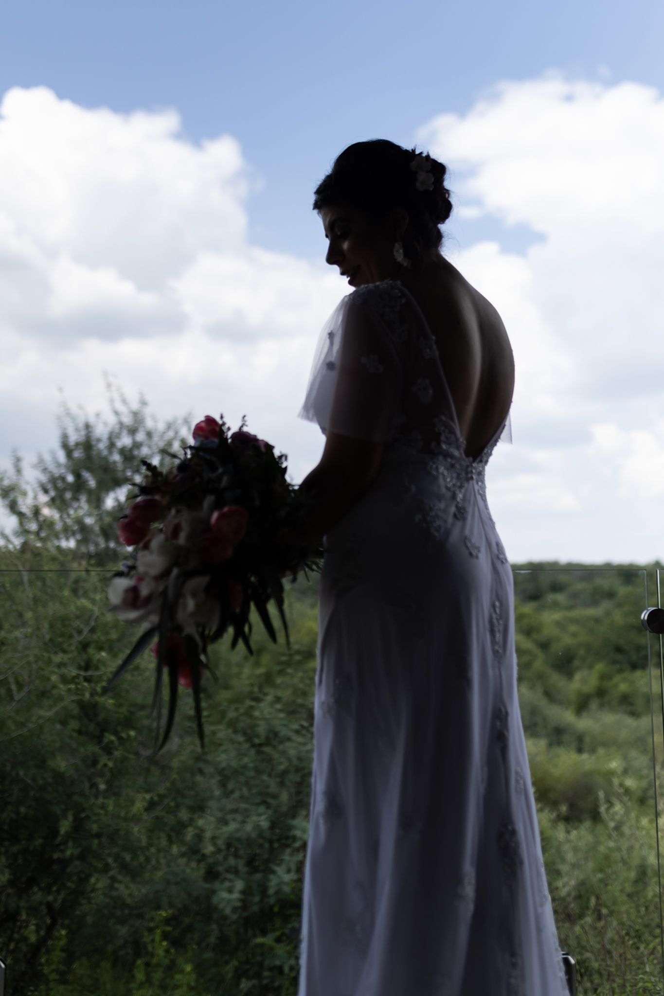 Silhouette of bride holding bouquet against cloudy sky shows dramatic backlit wedding portrait.