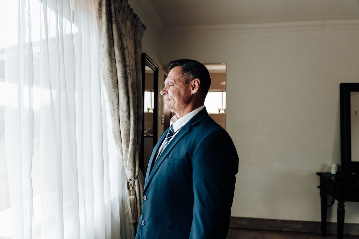 Man in navy suit gazes thoughtfully out window in preparation room.