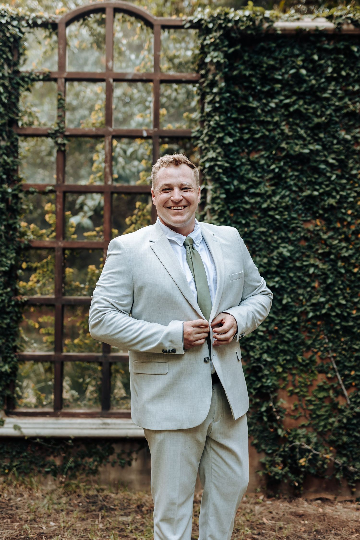 Groom adjusts his suit jacket while standing in front of an ivy-covered archway.