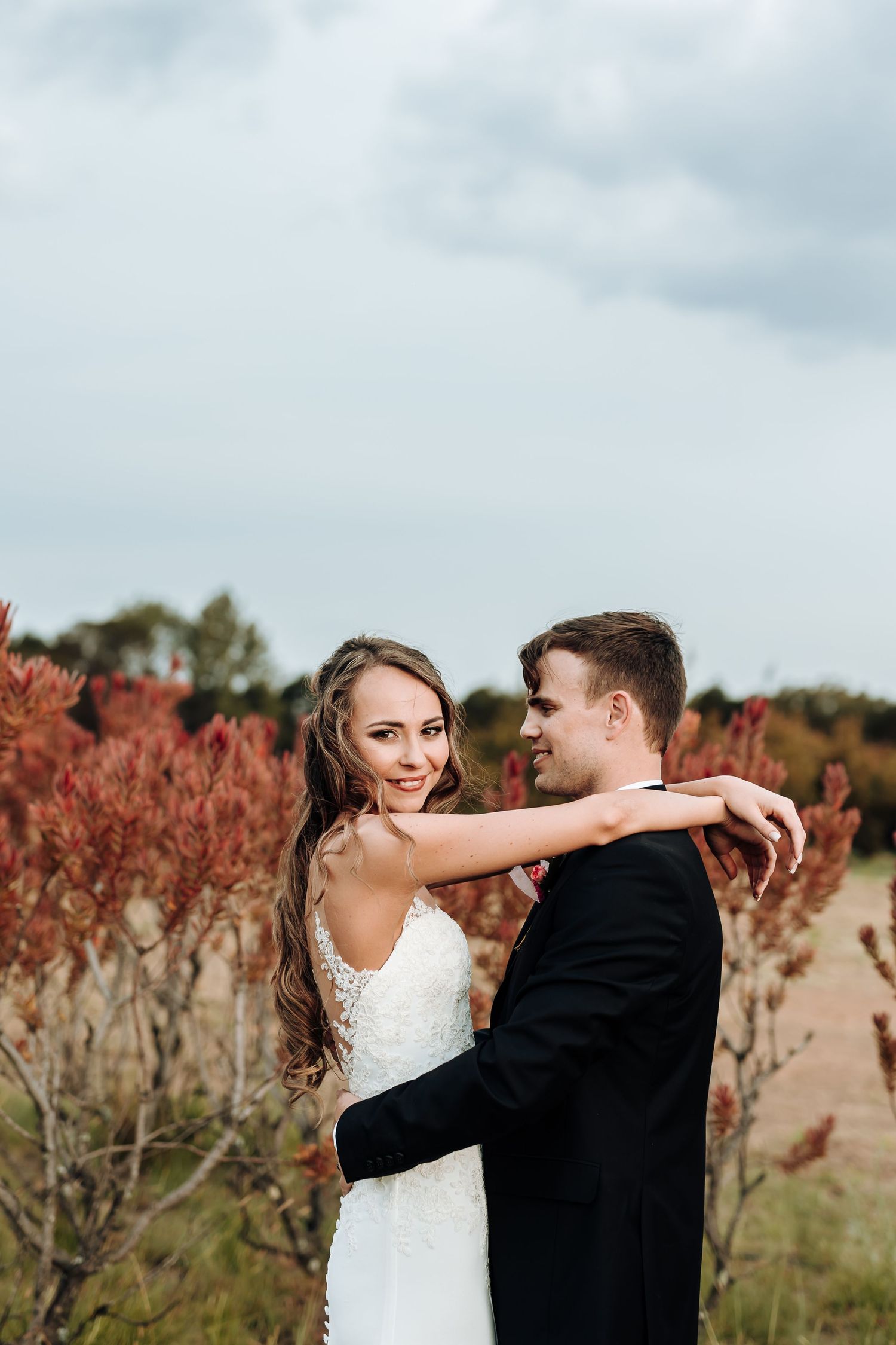 Bride in white dress embraces groom in black suit surrounded by red fall foliage.