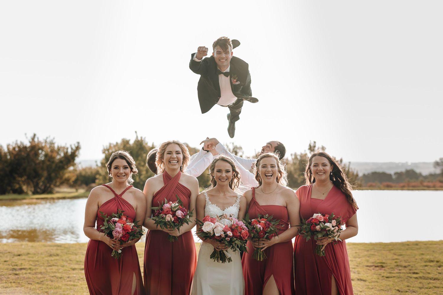 Wedding party poses outdoors by a lake as groomsman dramatically jumps in the background.