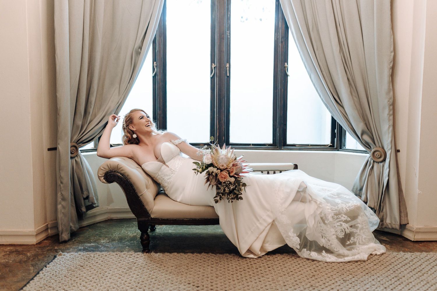 Bride in elegant gown poses on chaise lounge by draped windows in natural light.