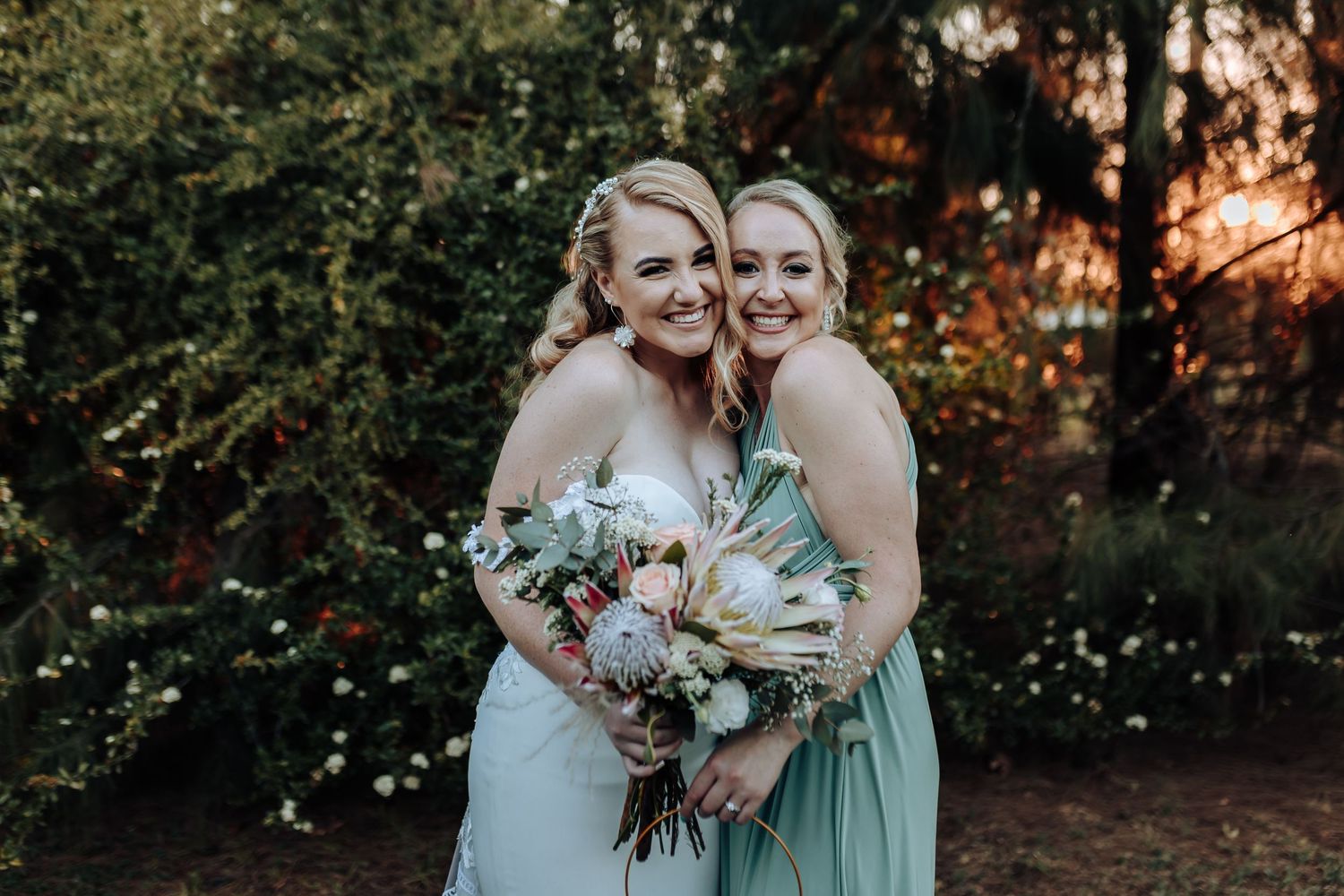 Two women in bridal party attire laugh together while holding elegant bouquets against natural outdoor backdrop.