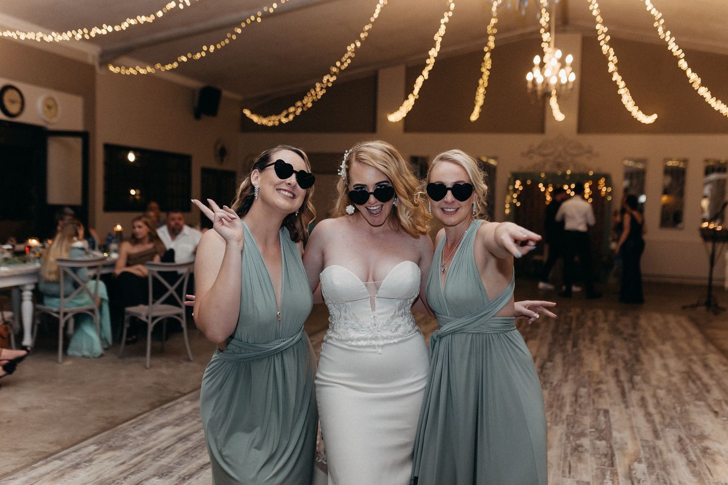 Three women wearing sunglasses pose playfully at a wedding reception under string lights.