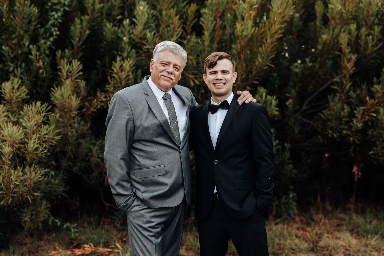 Two men in formal attire pose together against an evergreen backdrop in an outdoor portrait setting.
