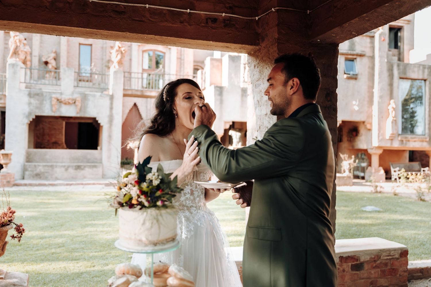 Bride and groom playfully feed each other wedding cake during their outdoor reception.