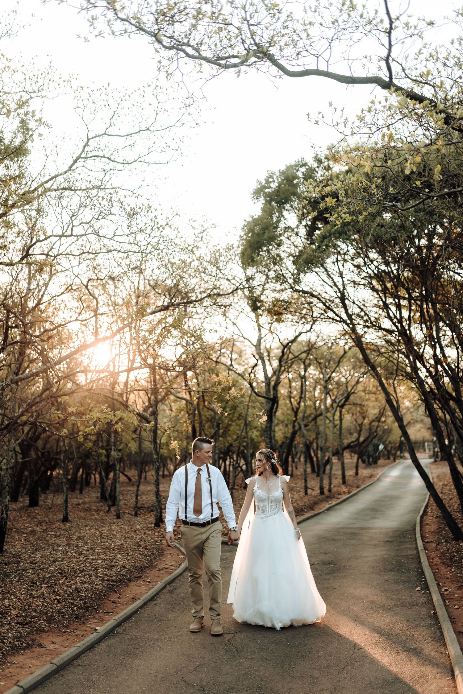 Newlyweds walking together on a tree-lined path at sunset.