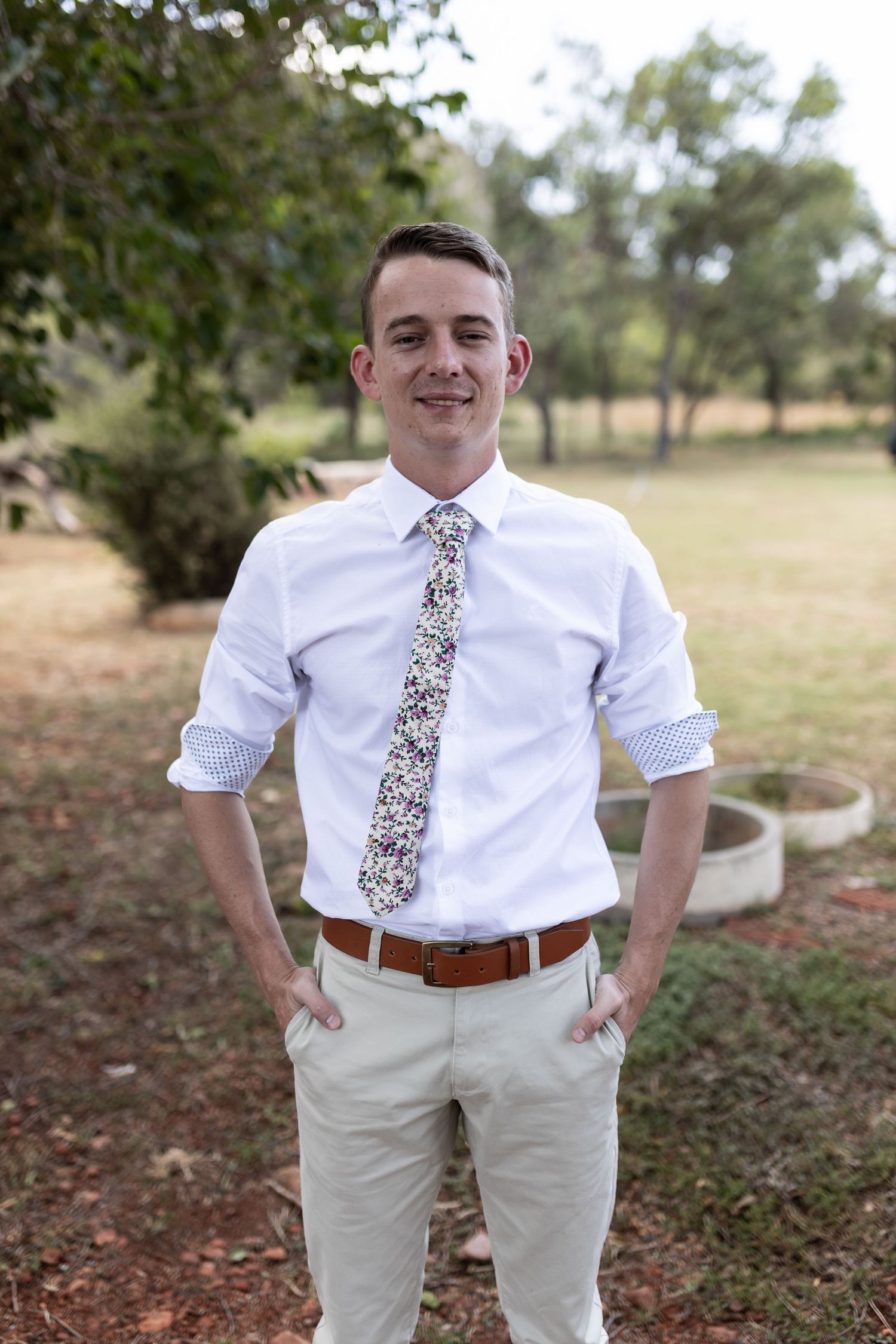 Groomsman in white dress shirt and floral tie poses outdoors in casual wedding attire.