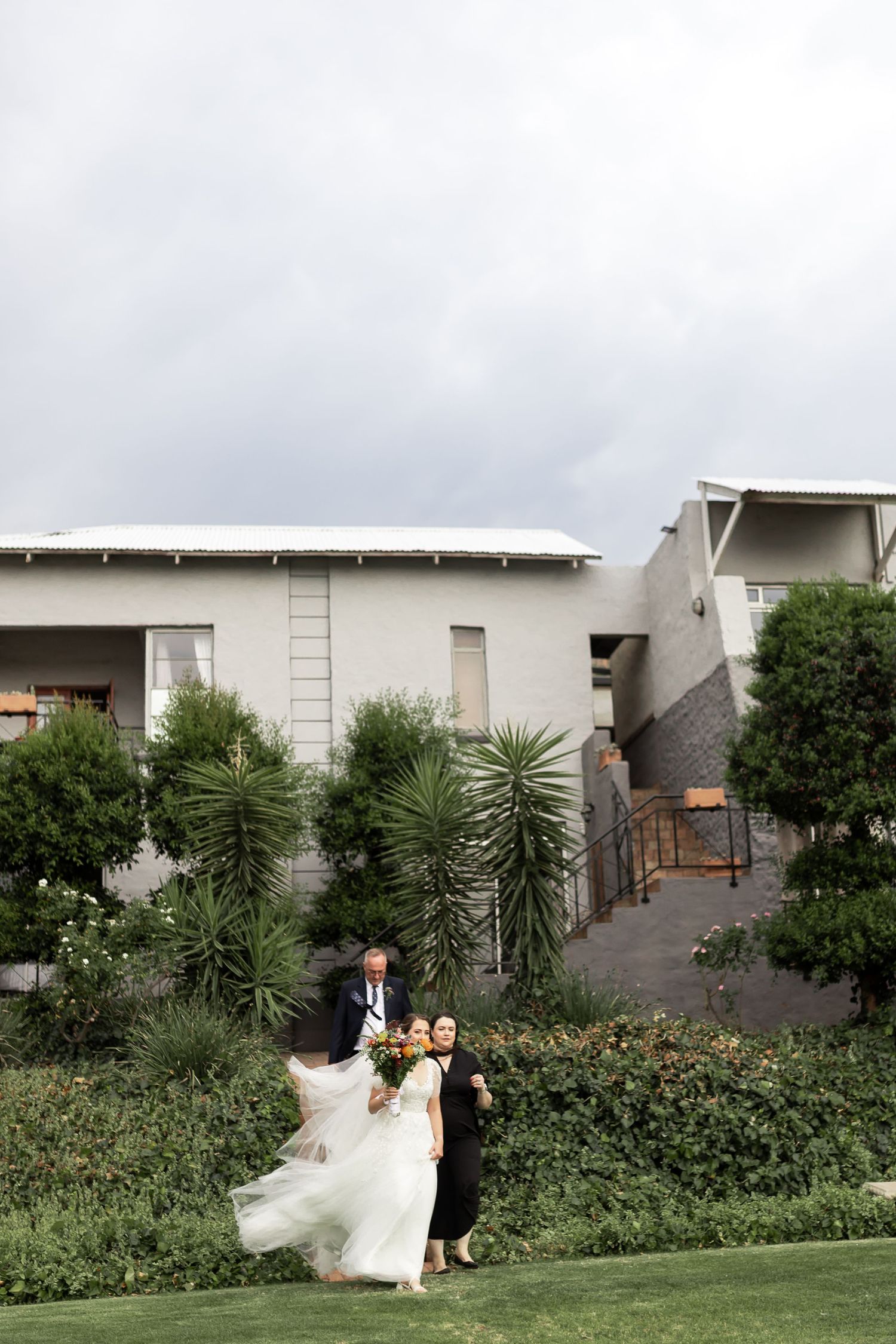 Dramatic wide shot of a wedding party descending a grassy hill with modern buildings behind them.