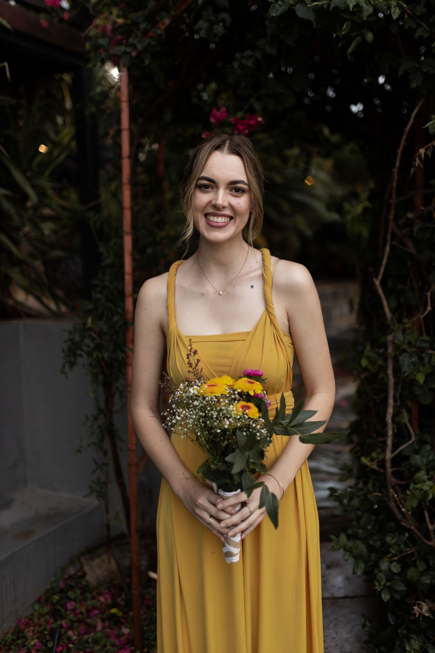 Bridesmaid in yellow dress holding rustic bouquet of wildflowers in garden setting.