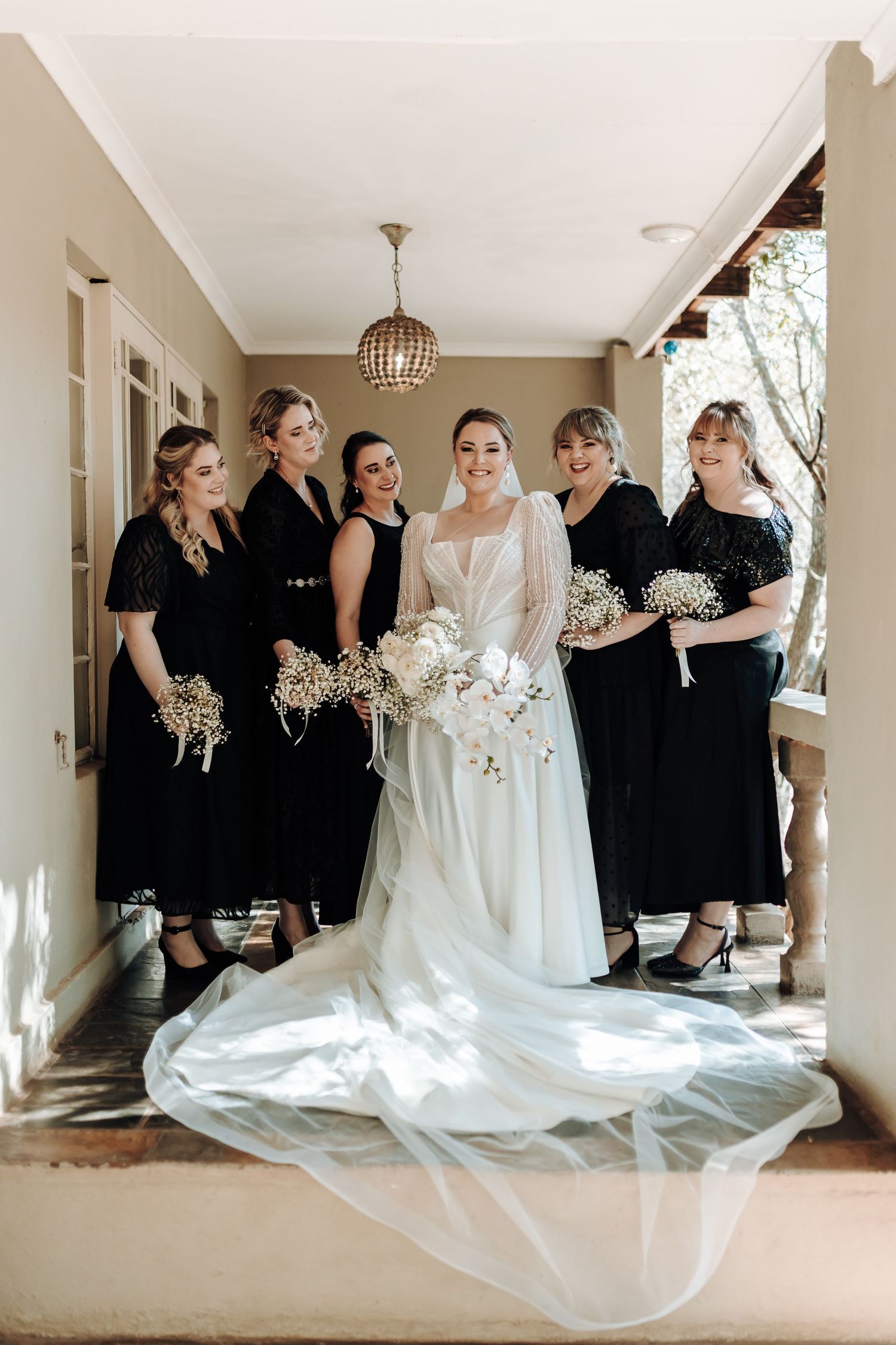 Wedding party poses in hallway with bride in white gown and bridesmaids in black dresses.