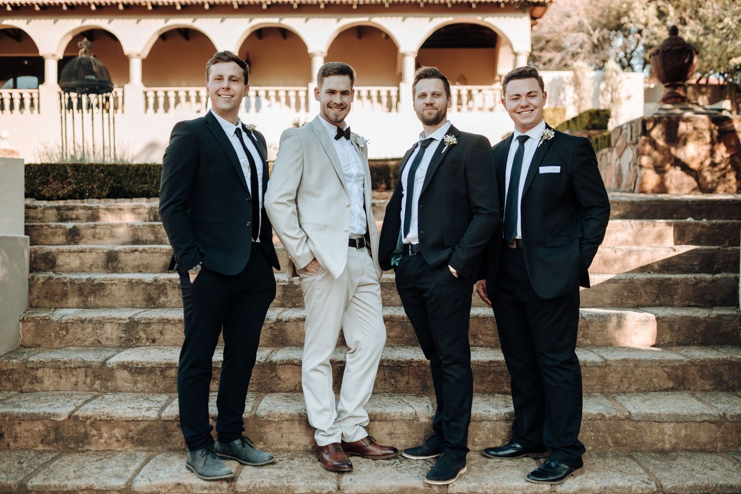 Four men in formal attire pose on stone steps with Spanish colonial architecture.