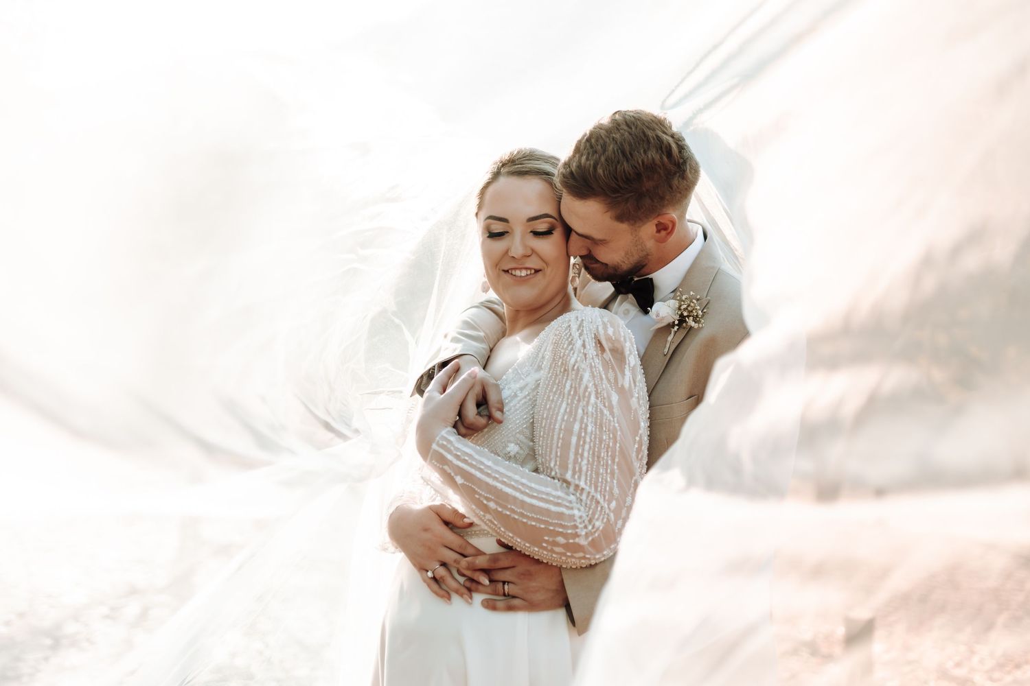 A romantic wedding couple embraces in soft lighting with flowing veil and beige suit details.