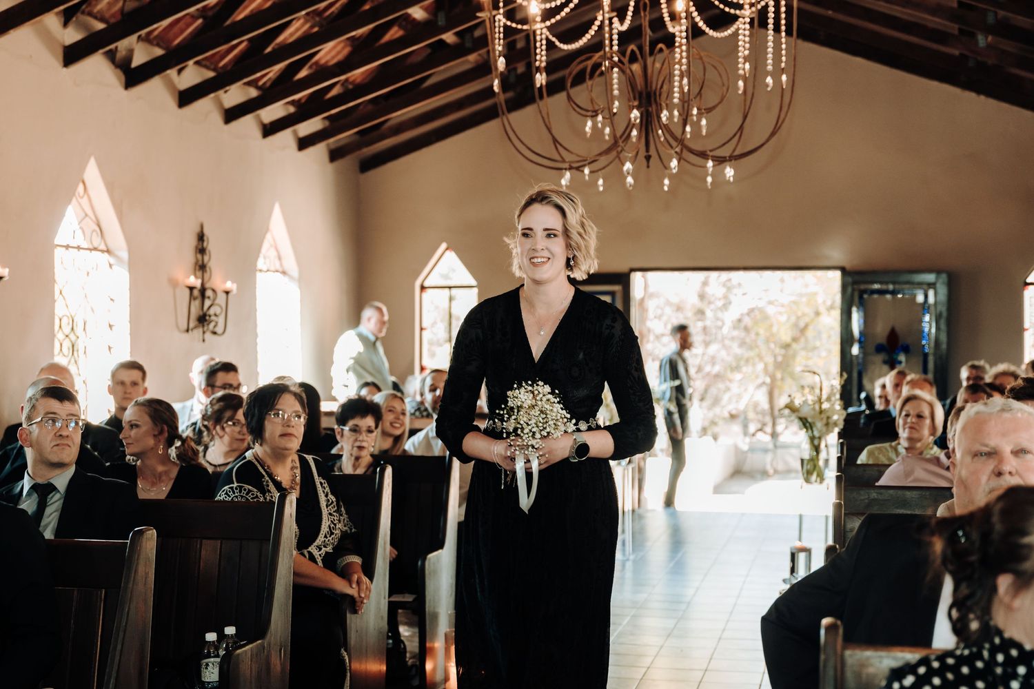 Person in black dress standing in church aisle during wedding ceremony with elegant chandelier overhead.