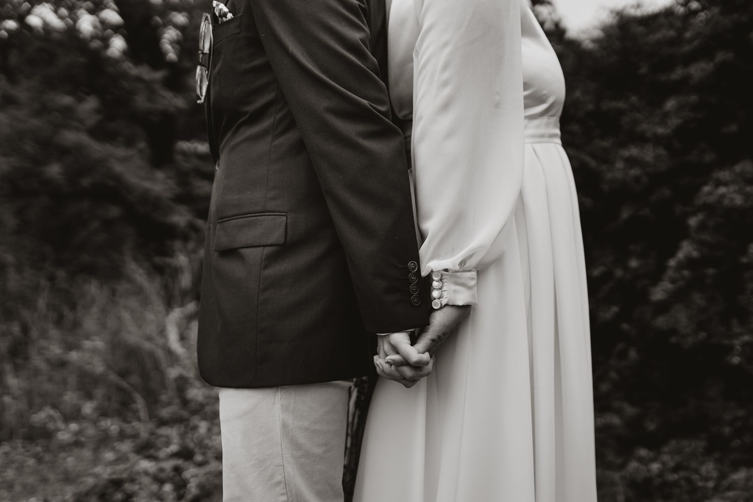 Black and white photo showing a close-up of a couple holding hands behind their backs on their wedding day.