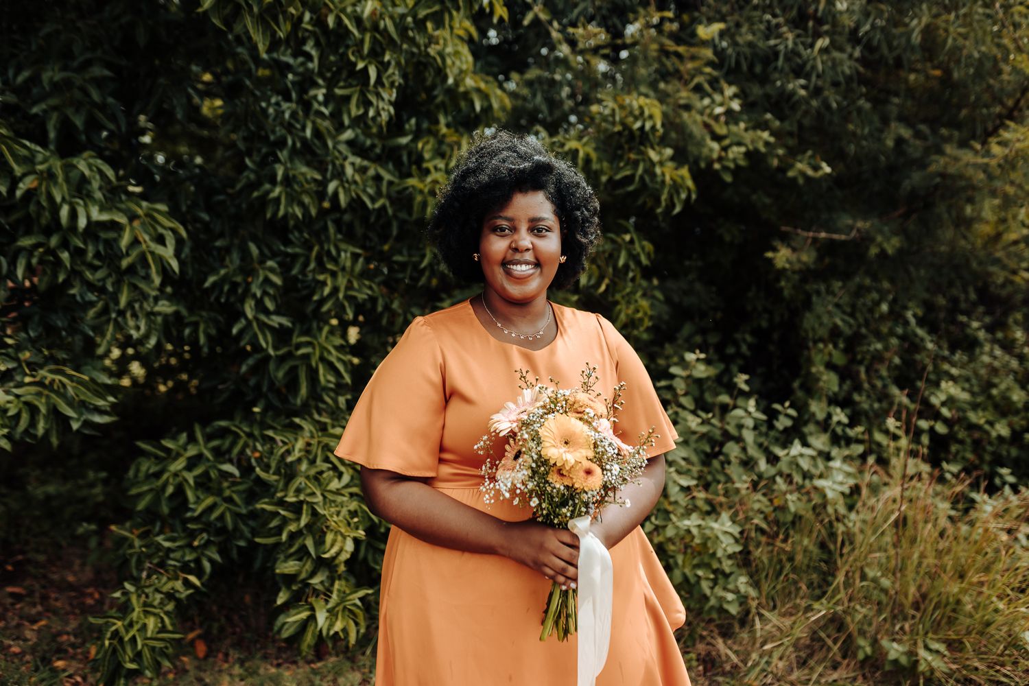 Person in orange dress holding peach and cream colored bouquet outdoors.