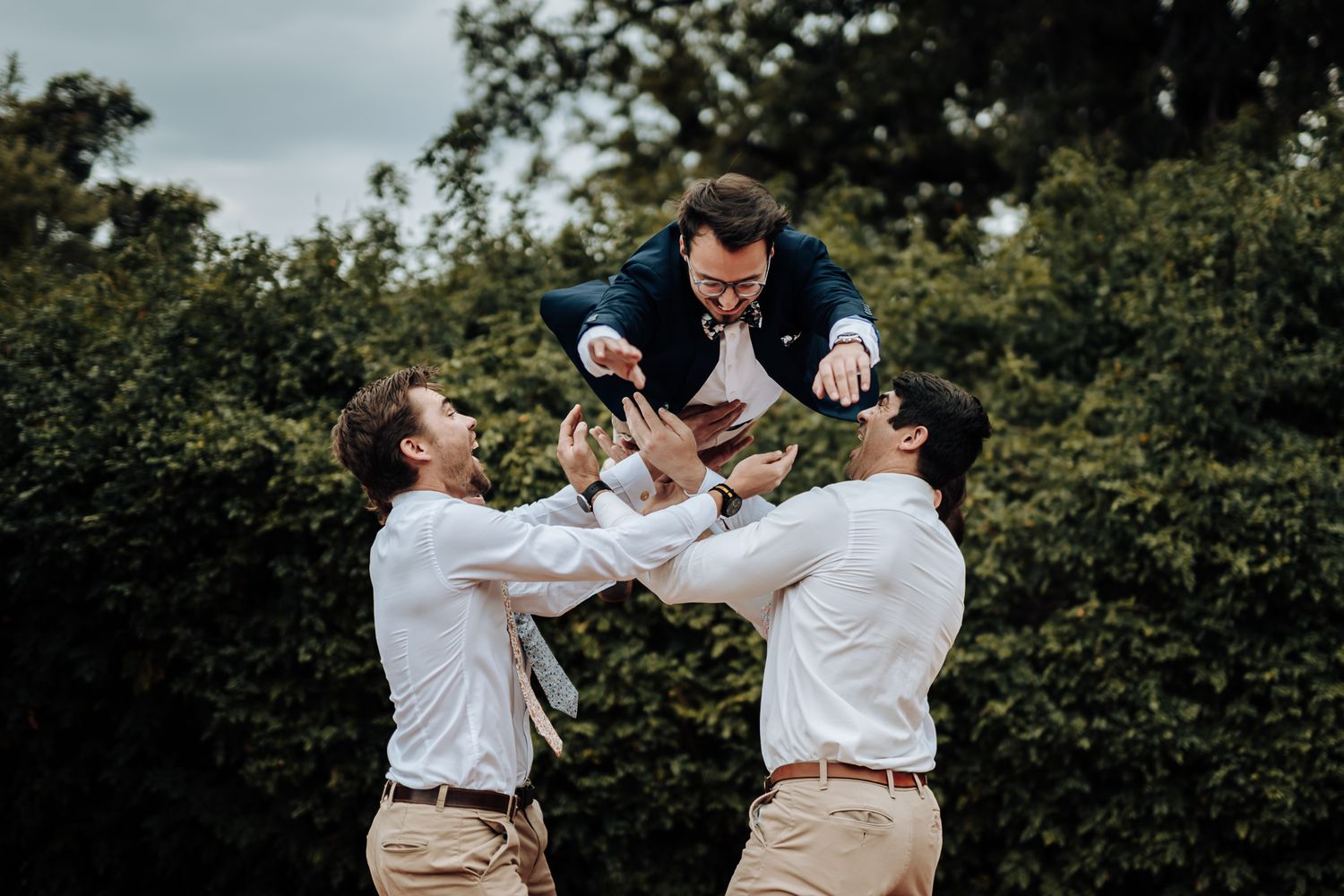 Groomsmen lifting groom in playful wedding party photo outdoors.