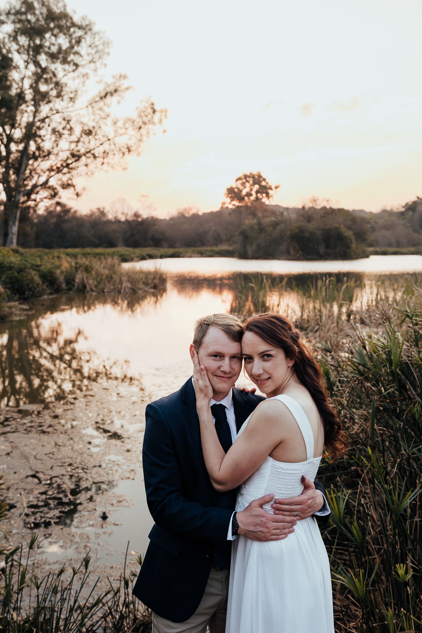 A tender embrace captured at golden hour beside a tranquil lake with reeds and natural landscape.