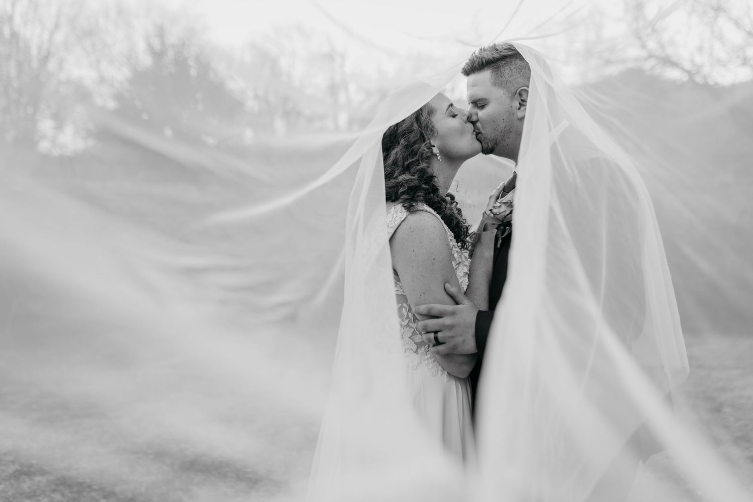 A romantic wedding couple embraces under a flowing white veil in a black and white photograph.