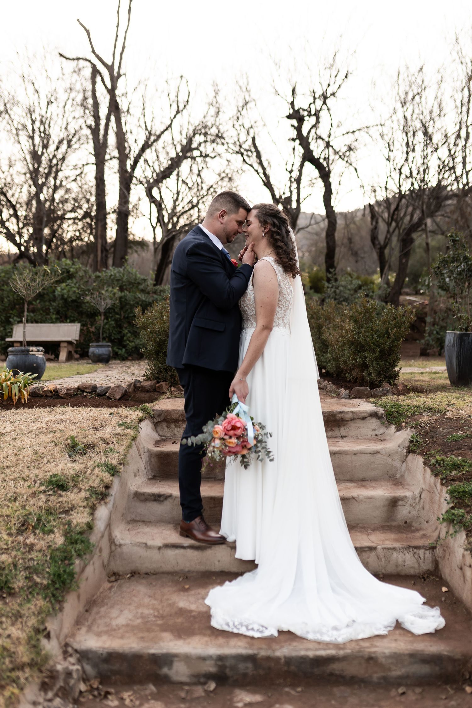 A romantic wedding couple sitting on stone steps in a natural outdoor setting with bare trees in the background.