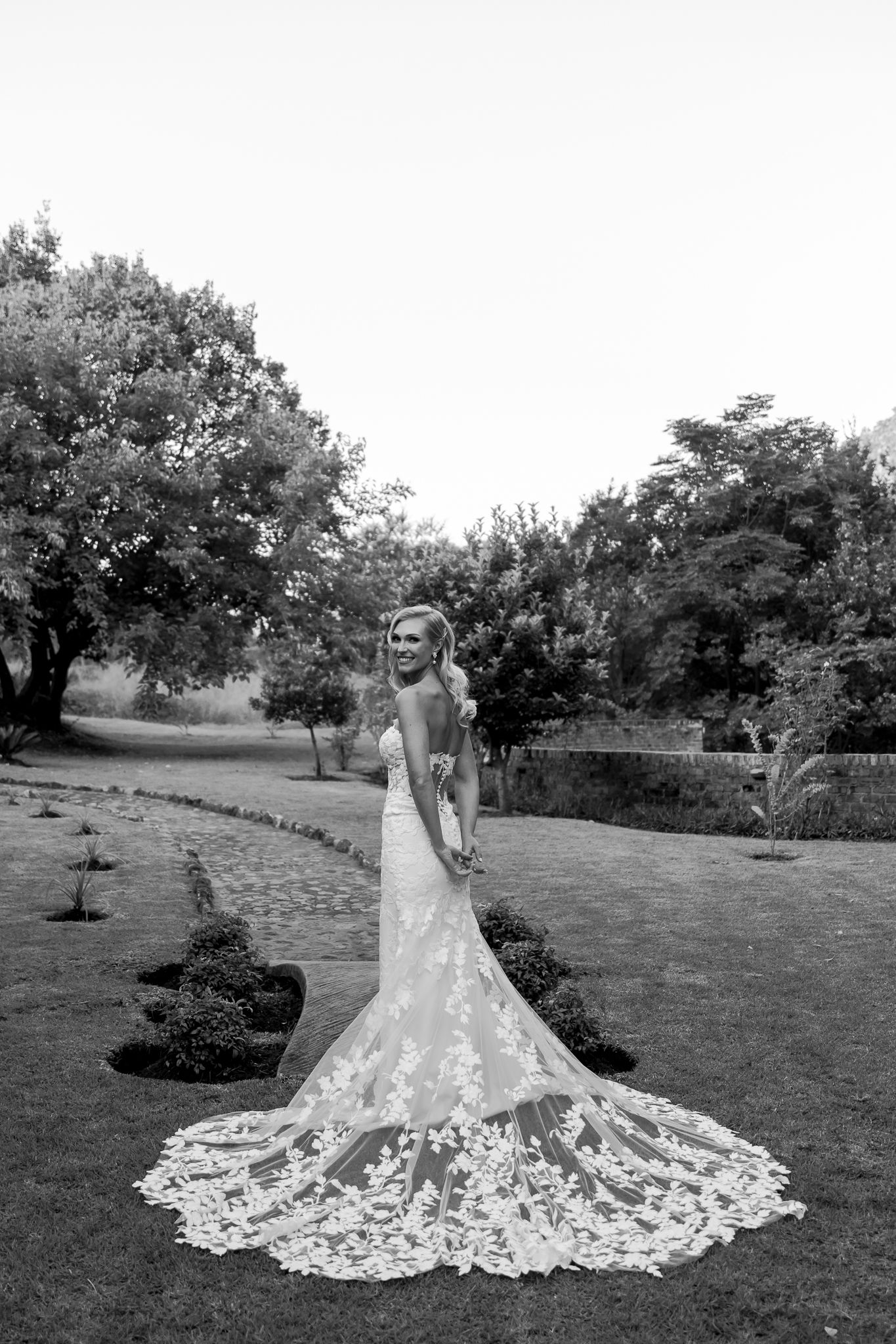 Black and white bridal portrait showcasing detailed lace train of wedding gown on manicured lawn.