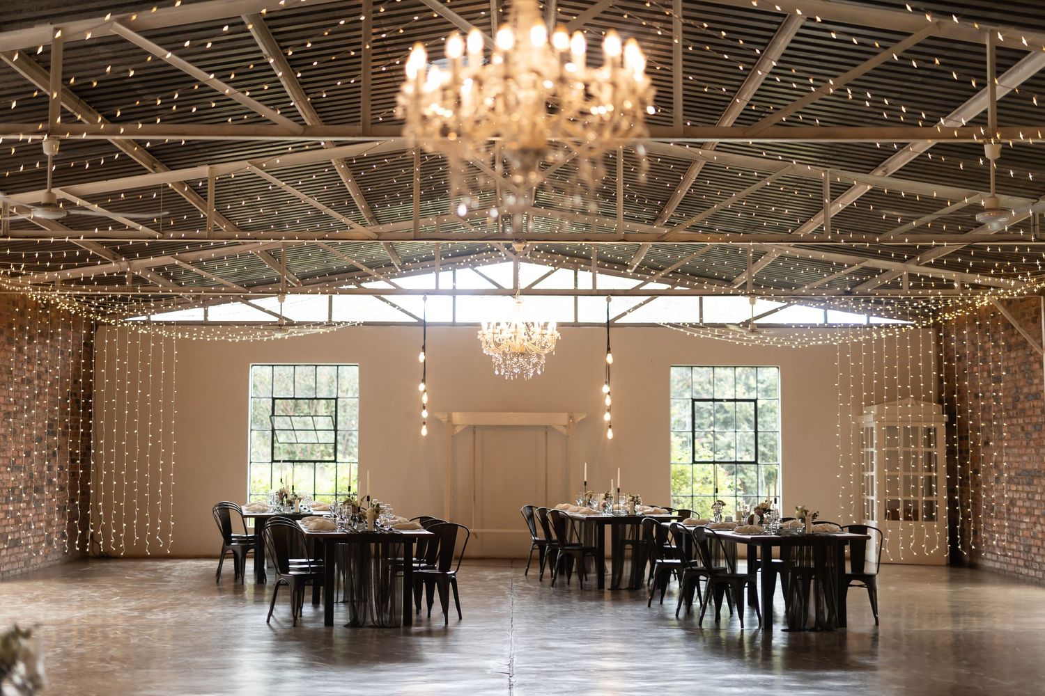 Elegant wedding venue with chandelier and string lights illuminating tables set for reception in white modern space.