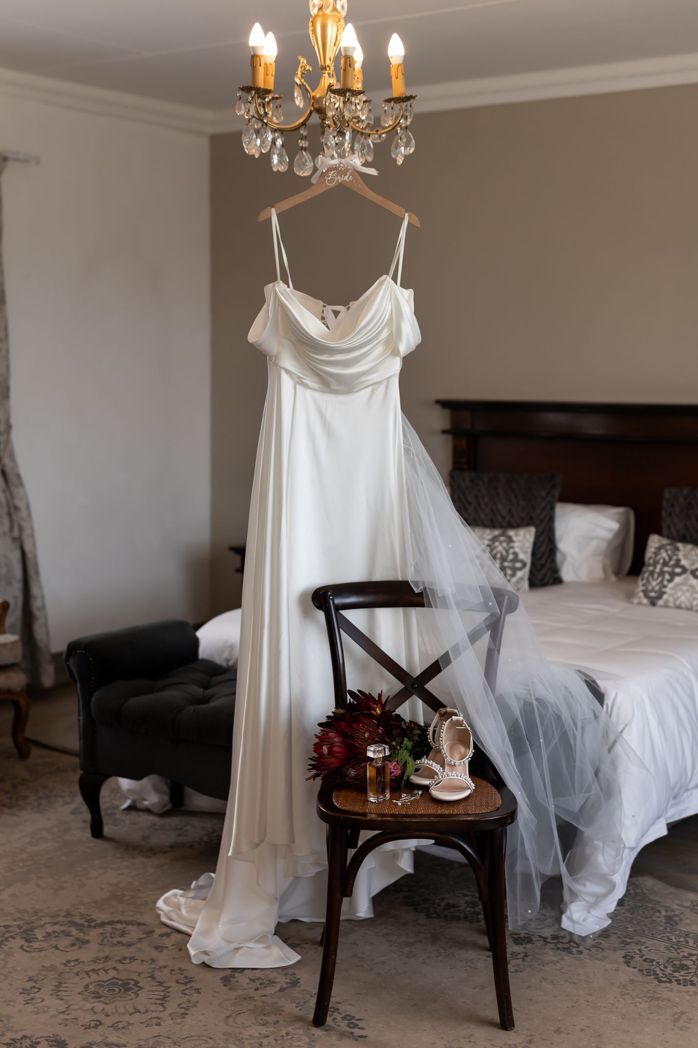 A white wedding dress hangs on a wooden hanger beneath an elegant crystal chandelier in a bedroom setting.