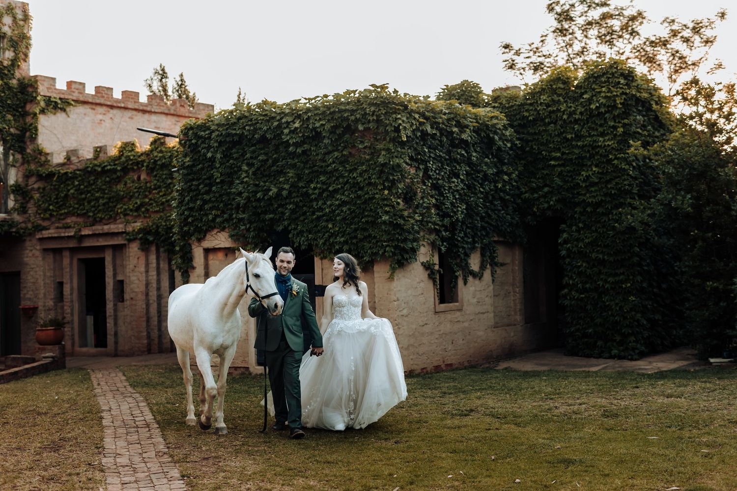 Newlyweds stand with a white horse on castle grounds surrounded by ivy-covered walls.