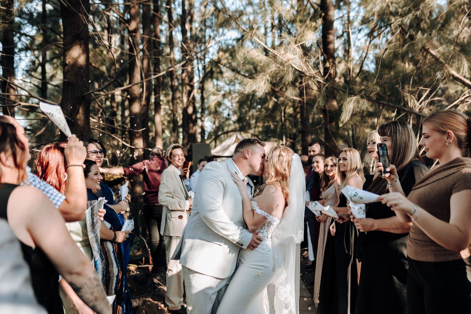 An intimate forest wedding ceremony with guests gathered around the couple during their first kiss.