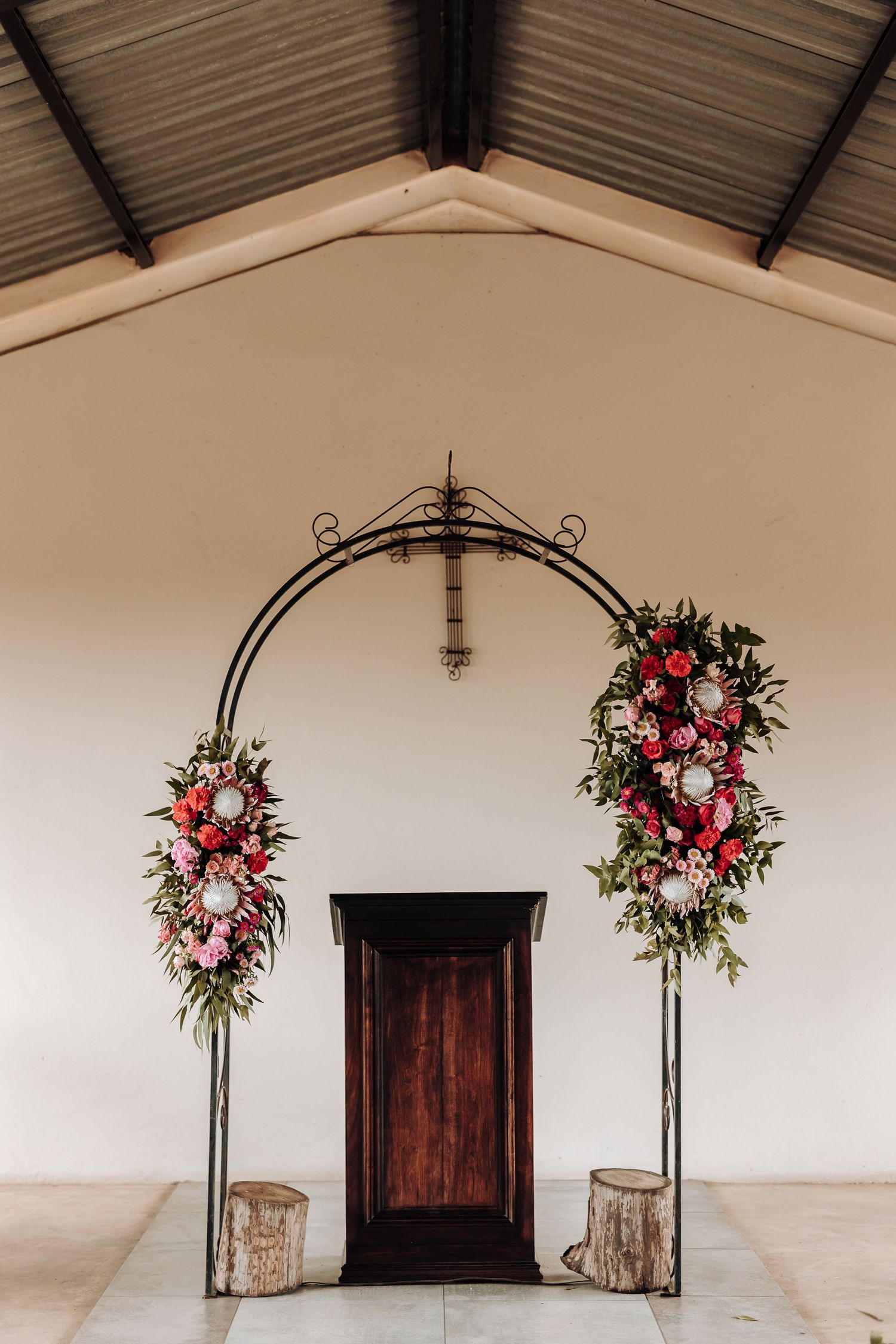 Metal wedding arch adorned with pink and white flowers framing a wooden door in a chapel.