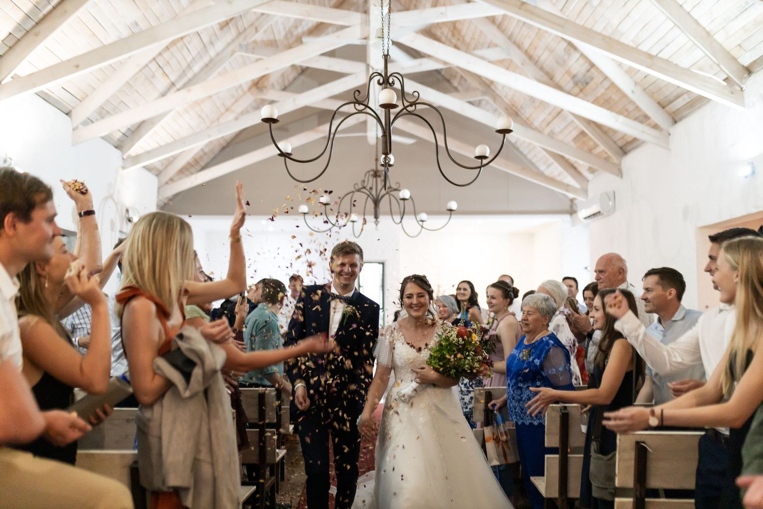 Wedding guests toss confetti at a celebration under a white vaulted ceiling with elegant chandelier.