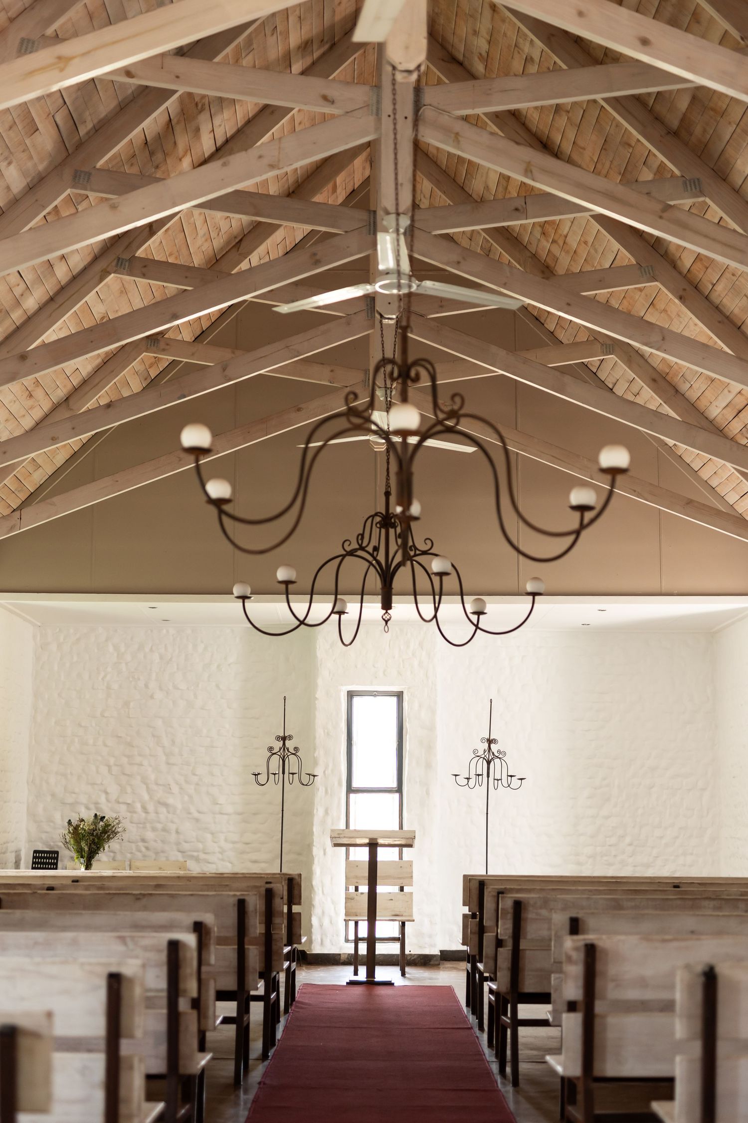 Interior of white chapel with wooden ceiling beams, chandelier, and wooden pews leading to altar.