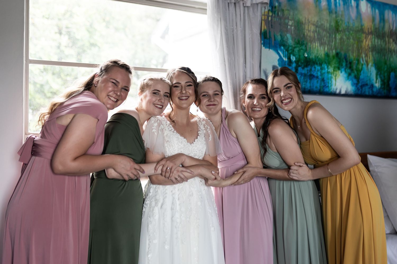 A bridal party in pastel dresses shares a group hug and laugh together.