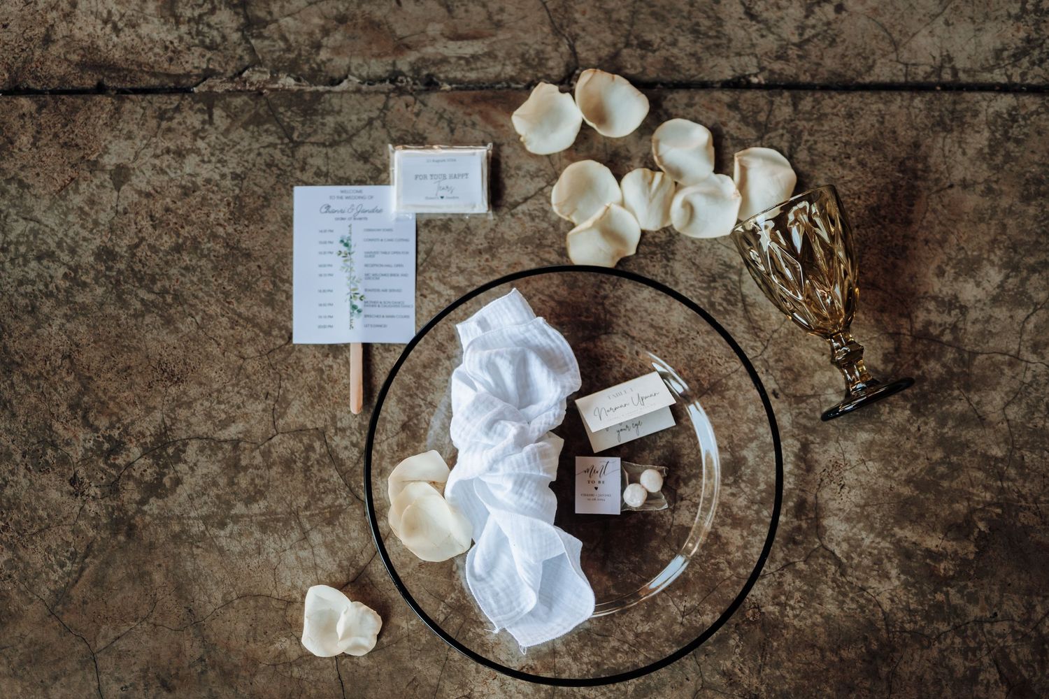 Wedding table place setting with black charger plate, napkins, and scattered white rose petals on rustic surface.