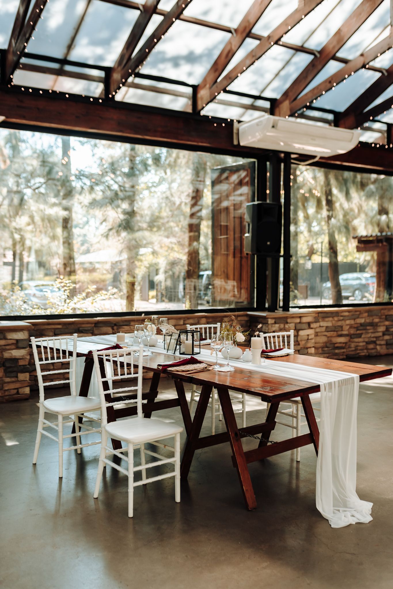 A rustic wedding reception table setup with white chairs and flowing fabric under a glass ceiling.
