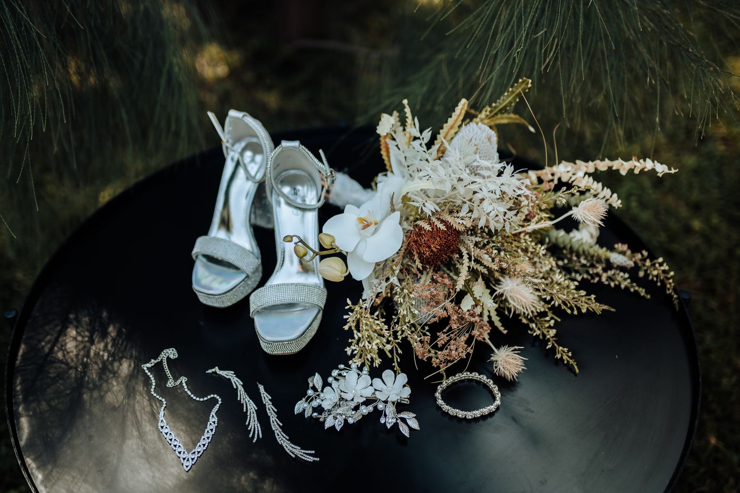 Wedding accessories including light blue heels, jewelry, and dried floral bouquet arranged on black surface.