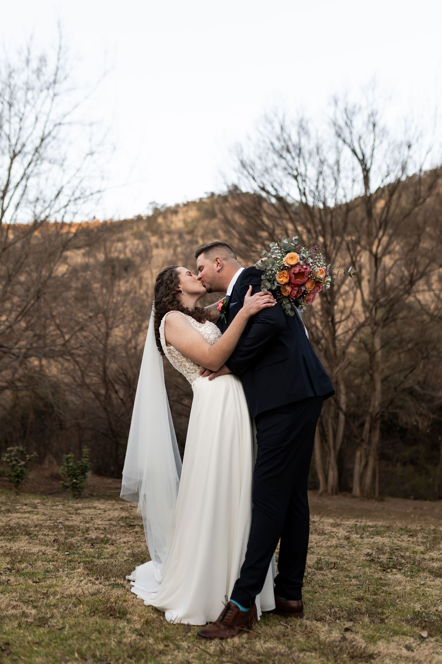 A couple shares a romantic kiss during their autumn outdoor wedding against a backdrop of bare trees and hills.
