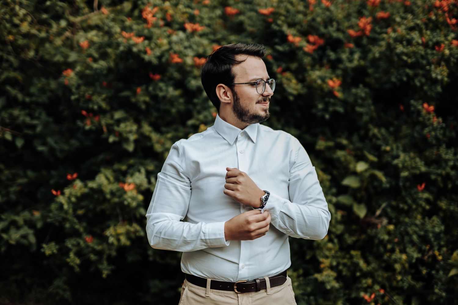 Elegant groom in white shirt and khakis adjusts sleeve against garden backdrop.