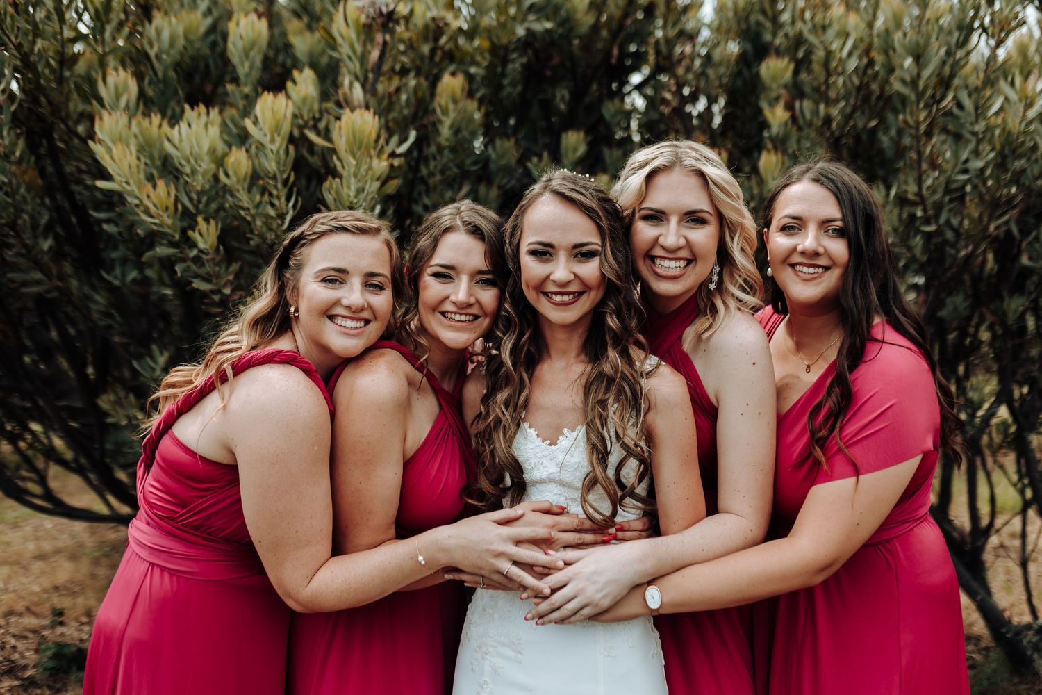 Bridal party in red dresses huddles close together for joyful group photo against green backdrop.