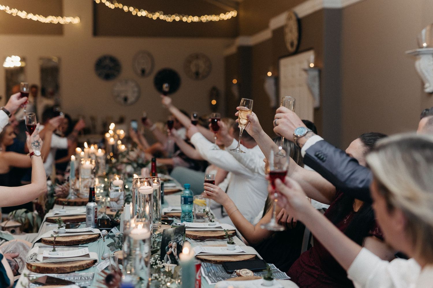 Wedding guests raising glasses for a toast at long reception table with glowing candlelight.