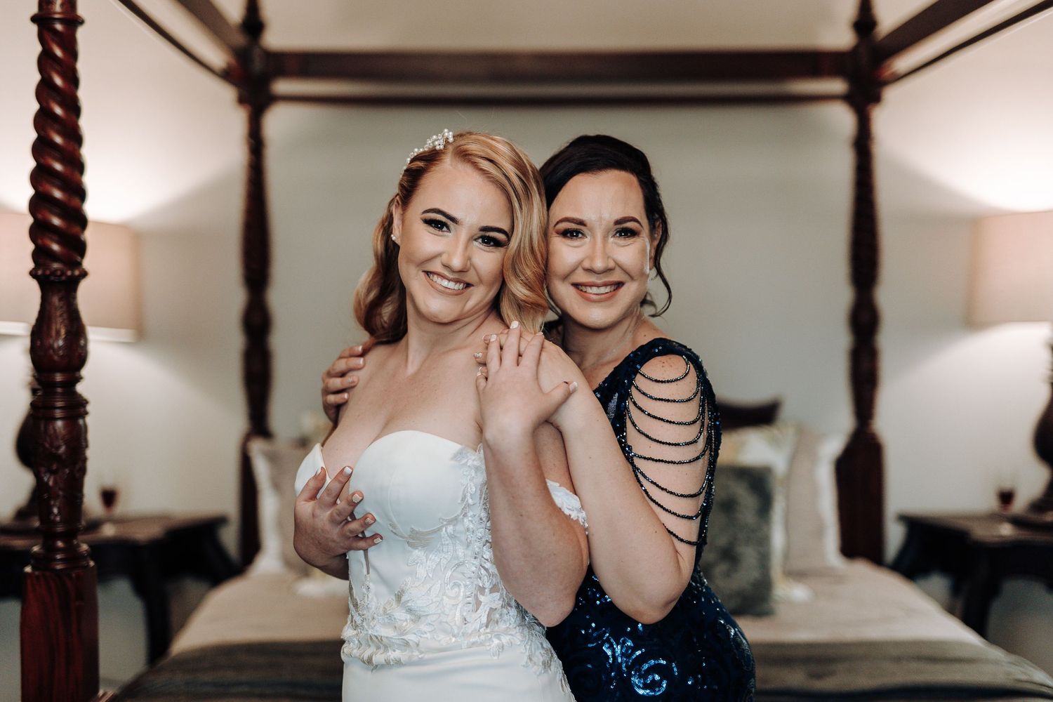 Wedding candid moment between bride and bridesmaid sharing joyful smiles by four-poster bed.