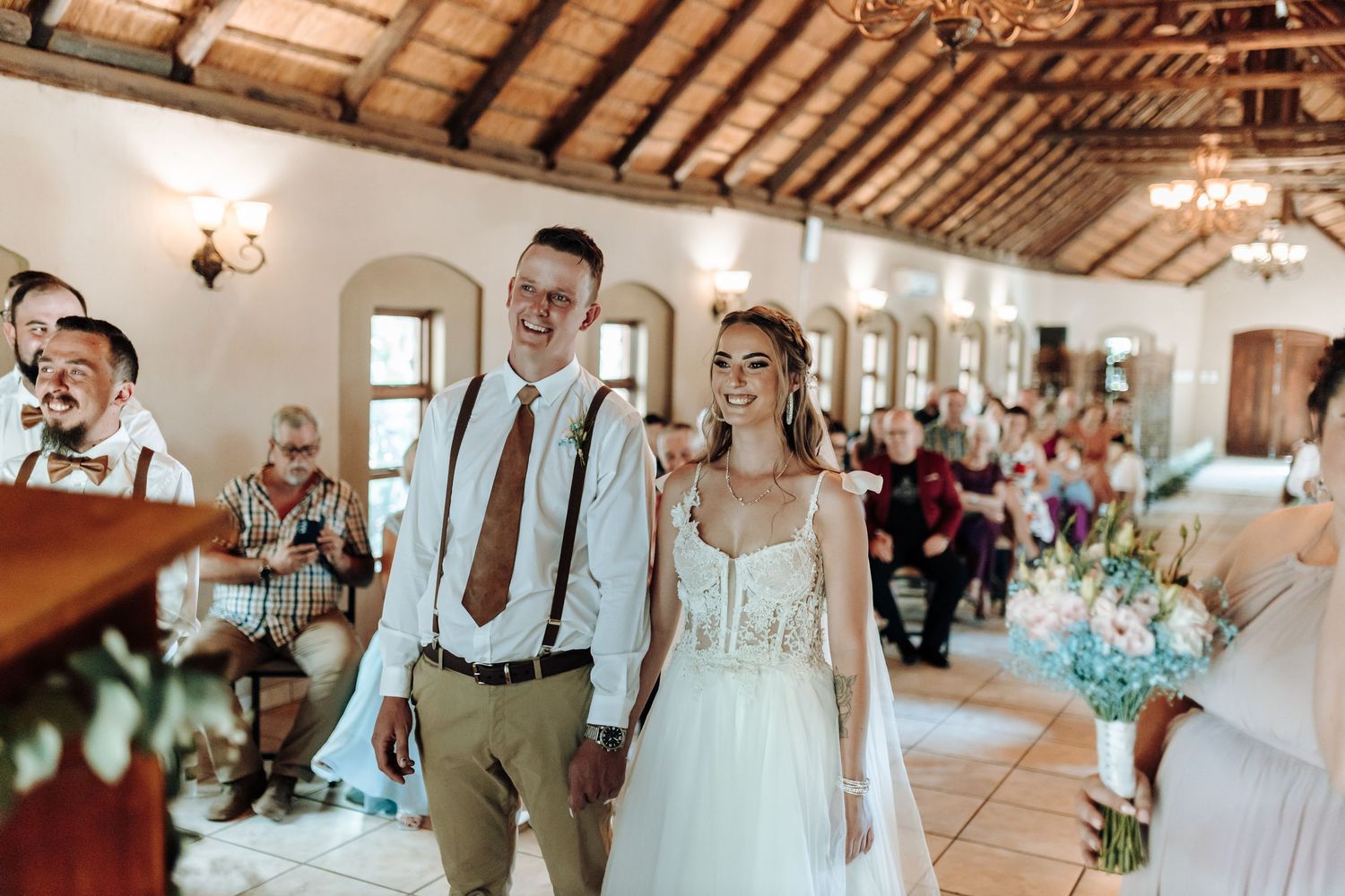 Couple walks down aisle in rustic venue with wooden beam ceiling and elegant chandeliers.
