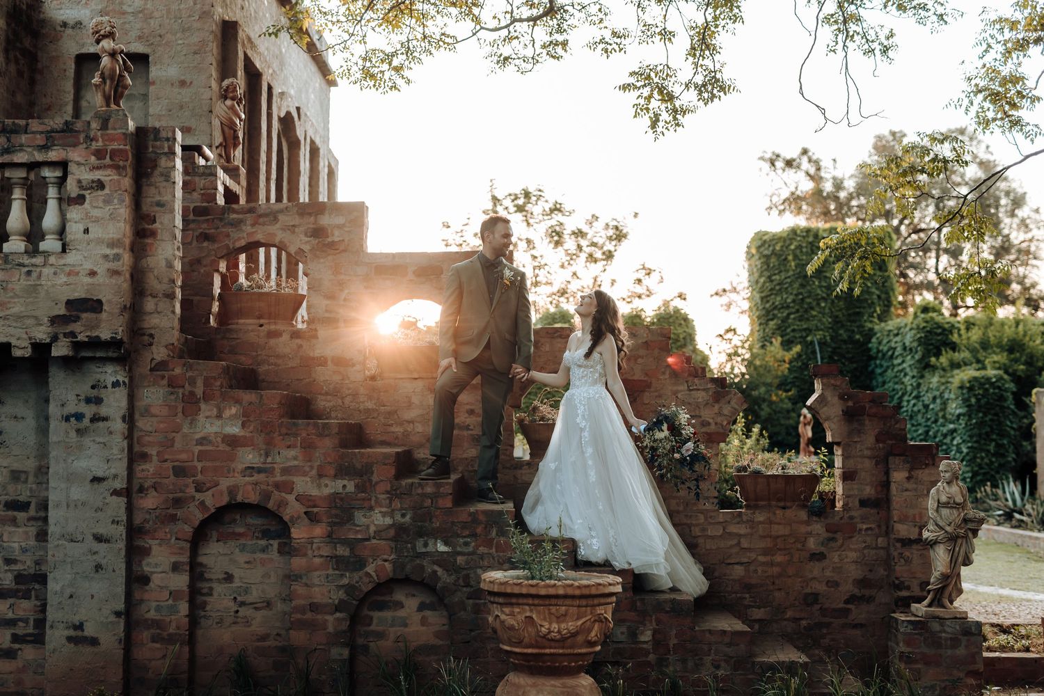 Bride in flowing gown stands atop castle steps at sunset with dramatic backlight.