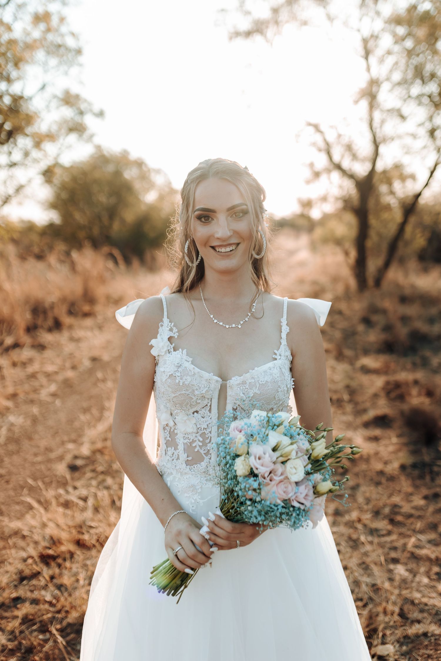 Bride in white lace gown holds pastel bouquet in golden sunset light.