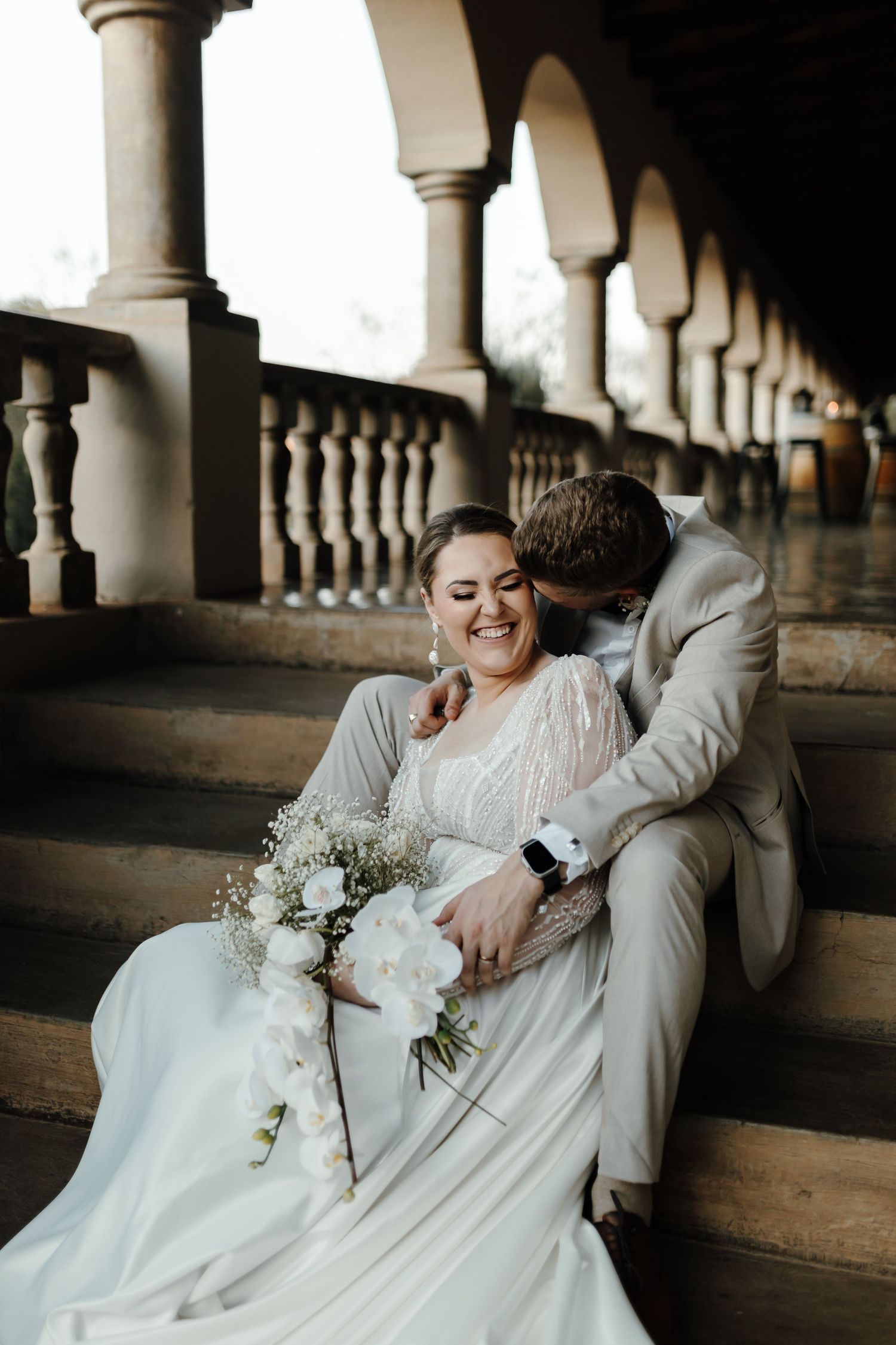 A romantic wedding moment captured under an arched colonnade with white pillars and balustrade.
