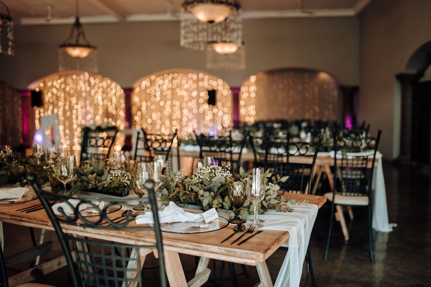Reception venue with wooden farm tables, black chairs, and dramatic fairy light backdrop.