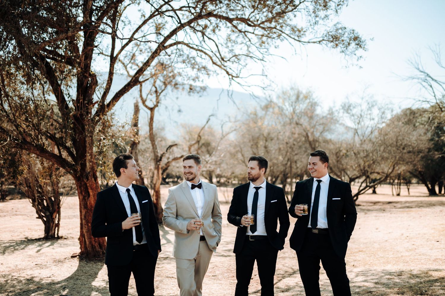 Groomsmen in formal attire stand together outdoors in a desert landscape with trees in the background.