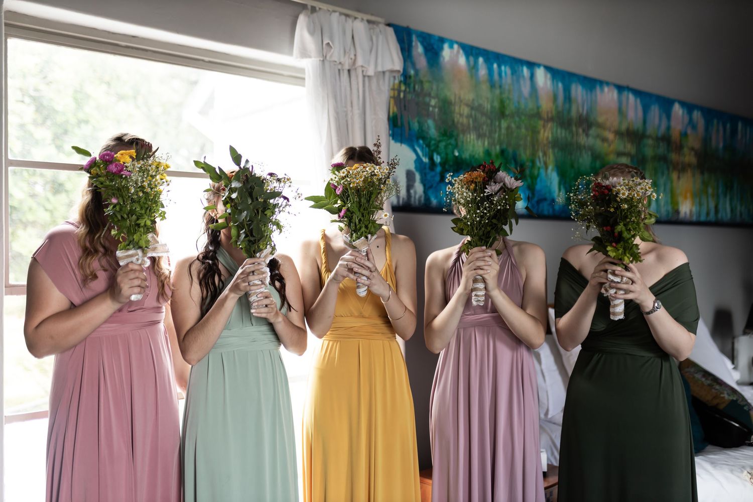 Five bridesmaids in pastel dresses holding floral bouquets in front of their faces.